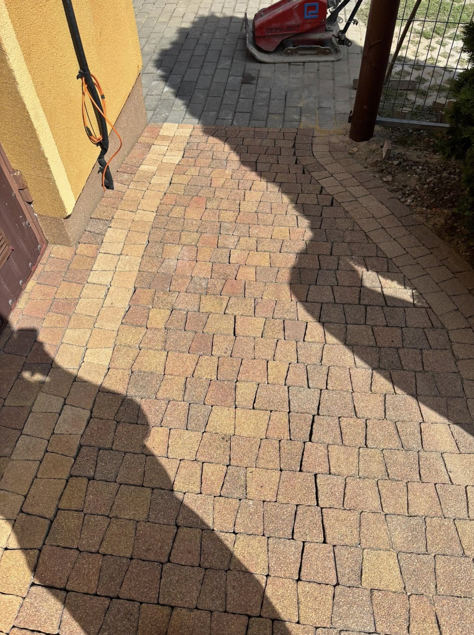 Ułożona kostka brukowa w odcieniach brązu i beżu, tworząca ścieżkę z delikatnym łukiem. W tle widoczna ubijarka i cień osoby.