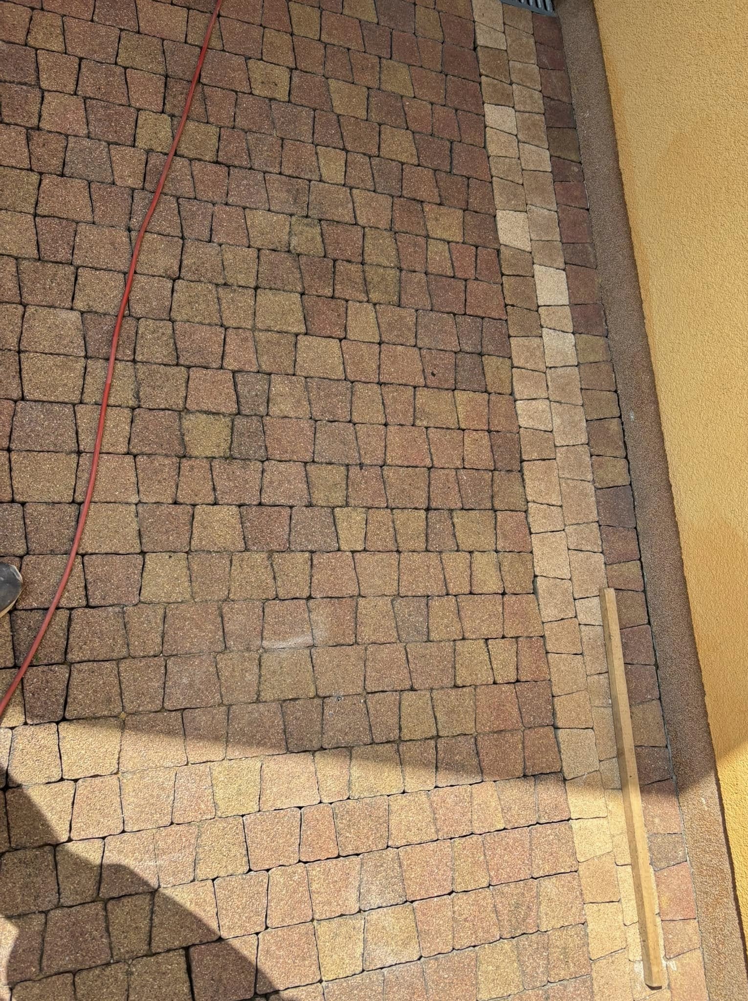 Ułożona kostka brukowa w odcieniach brązu i beżu, z widocznym kablem i fragmentem ściany w tle. Regularny wzór kostki, świeżo po ułożeniu, z detalami fug.