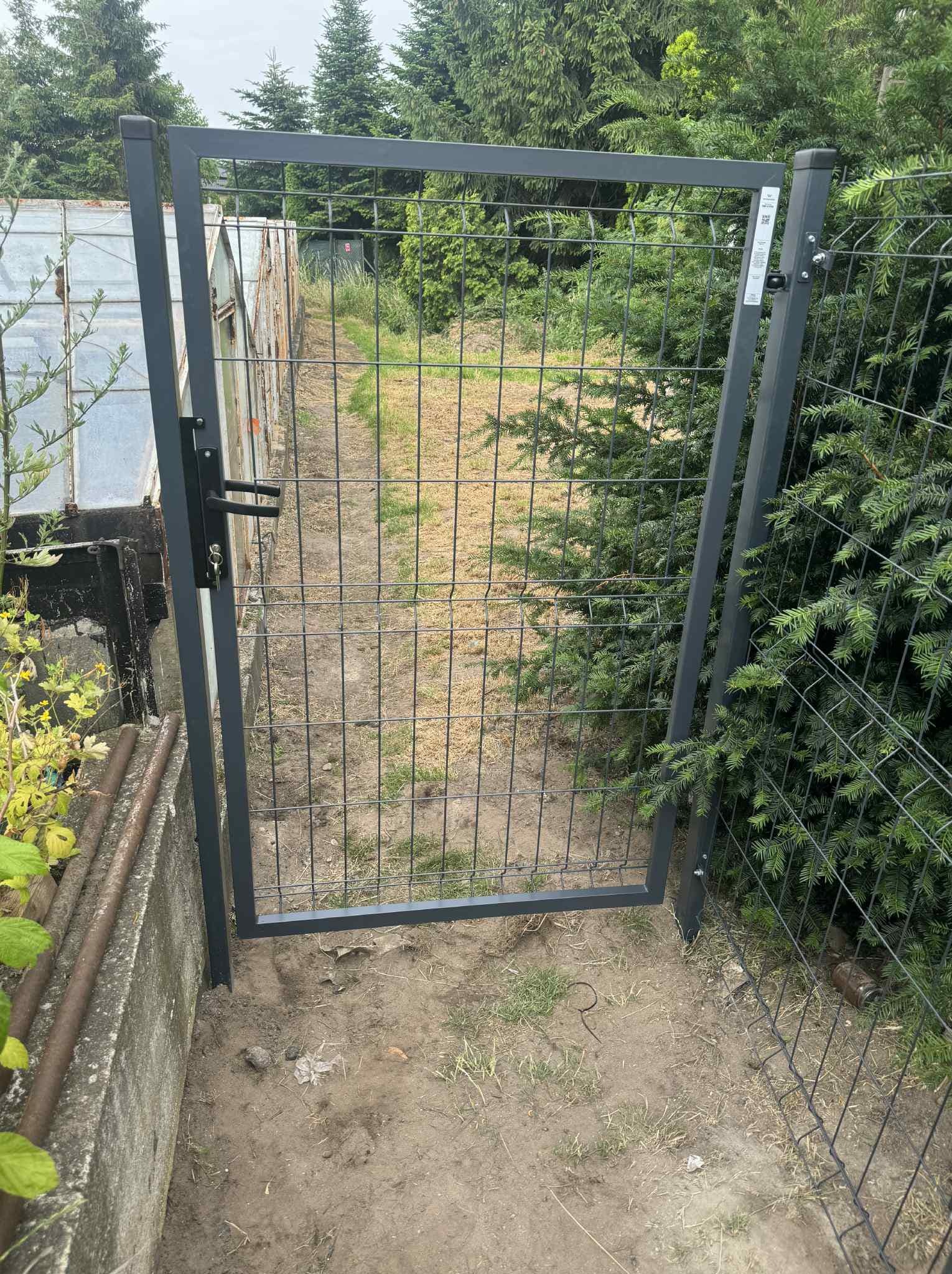 Szara, metalowa furtka z siatki, z czarną klamką i zamkiem, zamontowana w ogrodzie. W tle widoczny stary szklarniowy tunel i zieleń drzew.