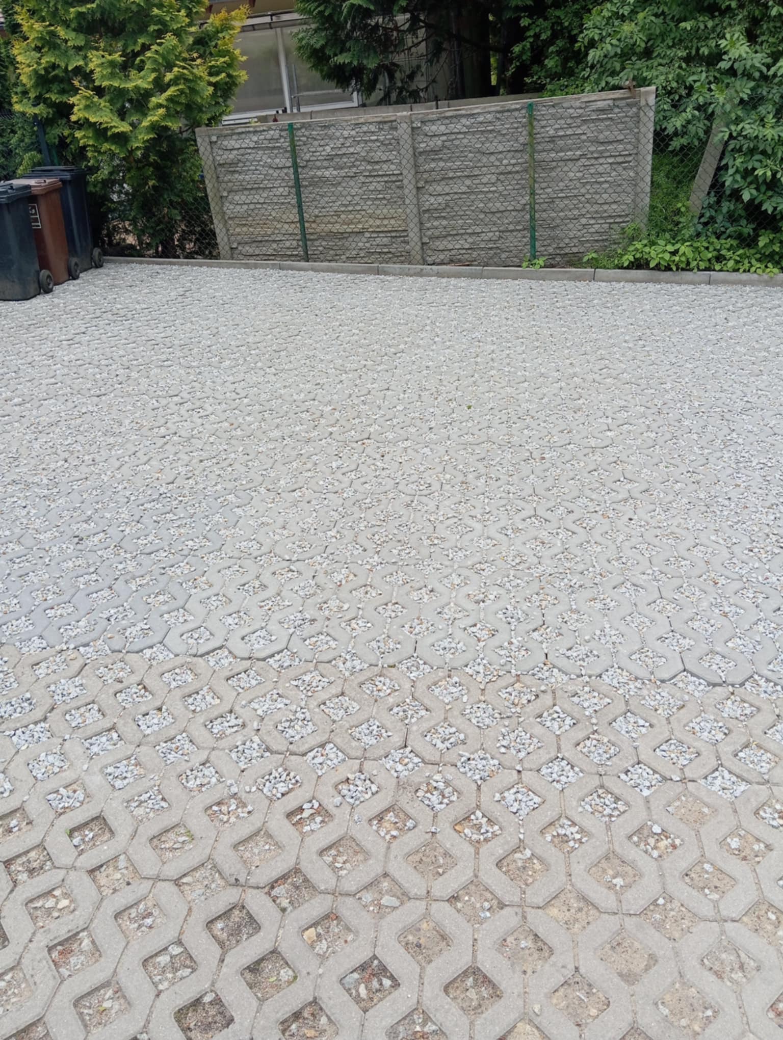 Szara ażurowa kostka brukowa wypełniona drobnym kamieniem na podjeździe, z widocznym fragmentem ogrodzenia i zieleni w tle. Widok z góry, szeroki plan.