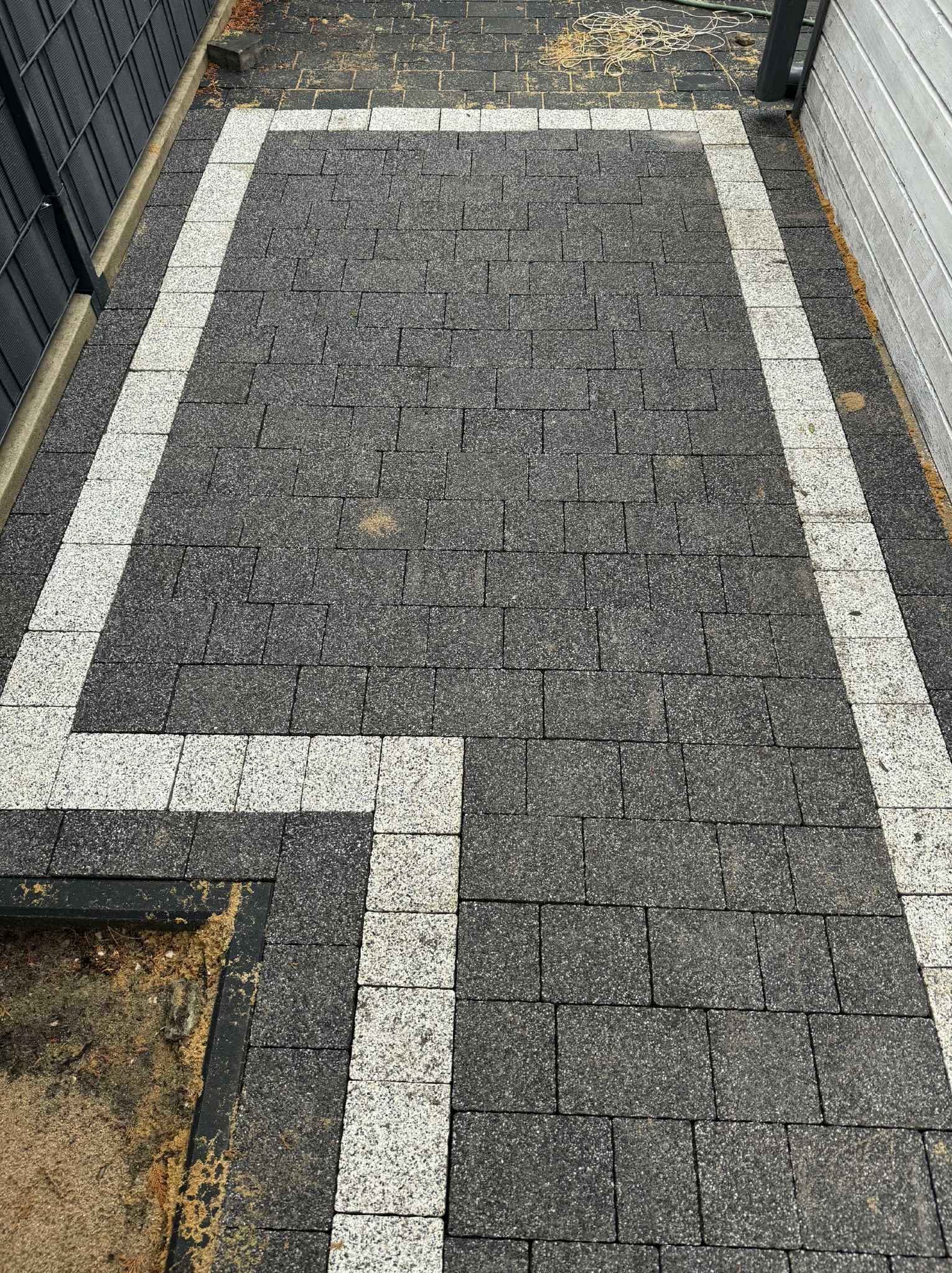 Ułożona kostka brukowa w odcieniach szarości z jasną obwódką. Widok z góry na ścieżkę z kostki, z wyraźnym wzorem i fakturą materiału.