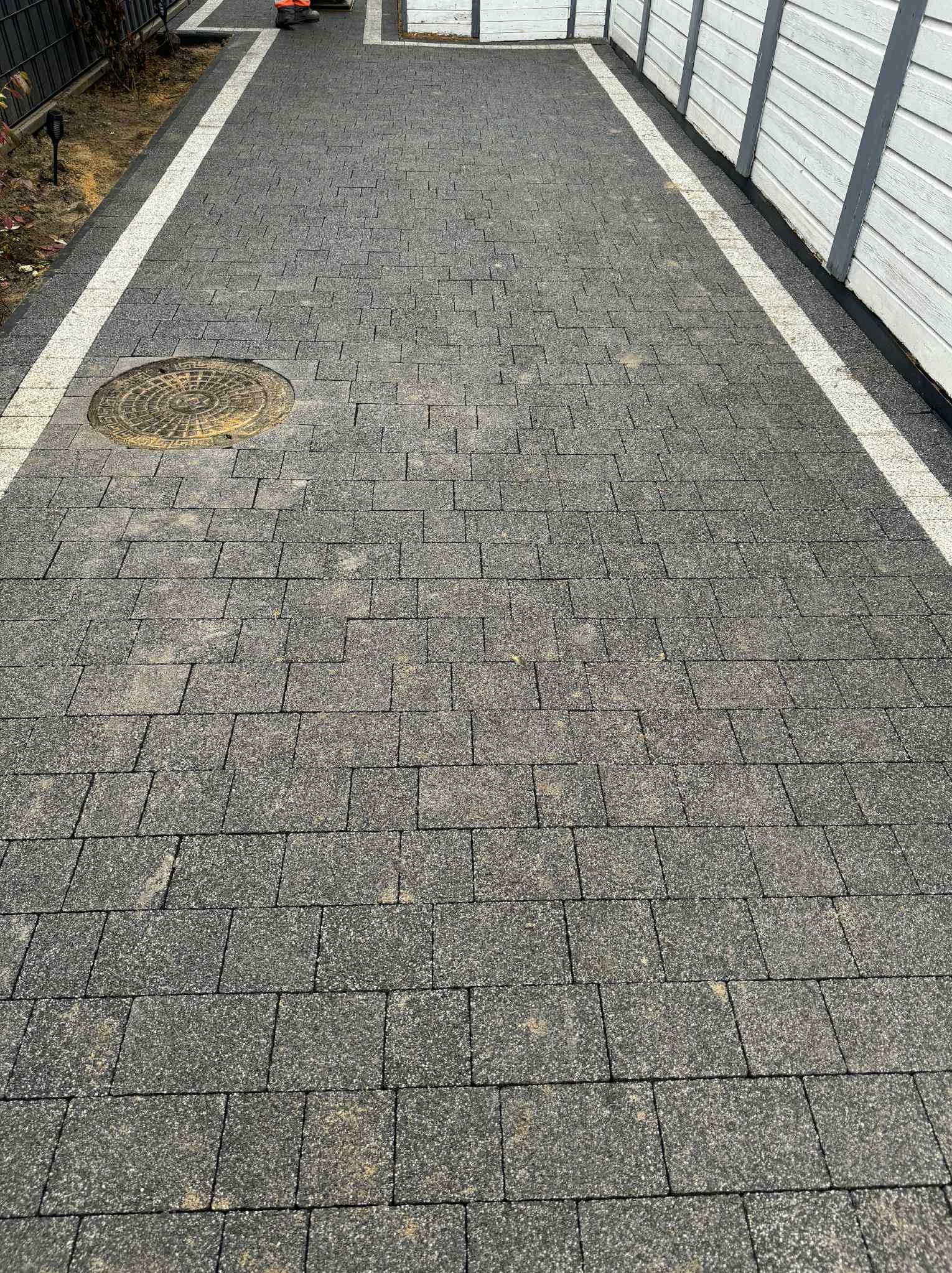 Ułożona, szara kostka brukowa z jasnymi obrzeżami i studzienką kanalizacyjną. Widok z góry na ścieżkę wzdłuż białego ogrodzenia.
