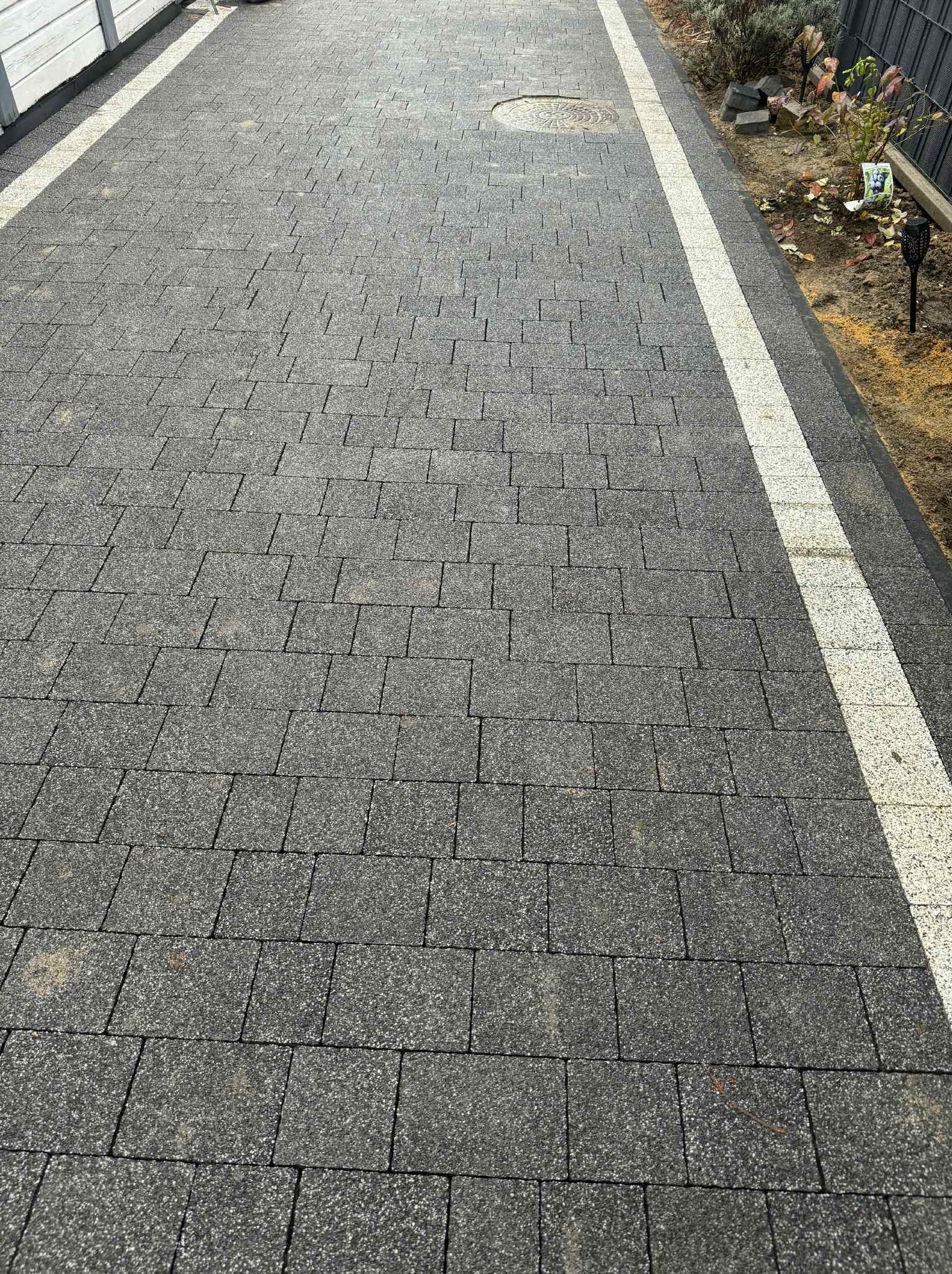 Ułożona szara kostka brukowa z białymi pasami po bokach, tworząca ścieżkę. Widoczny właz kanalizacyjny i fragment ogrodu z roślinami.