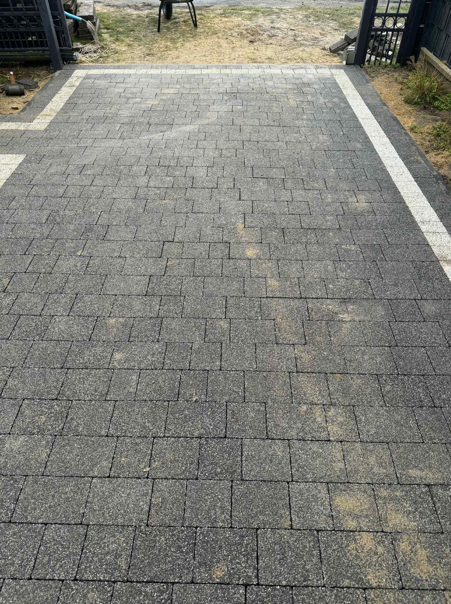 Szara kostka brukowa ułożona wzdłuż ścieżki z białymi obrzeżami, widok z góry, z elementami zieleni po bokach i fragmentem bramy w tle.