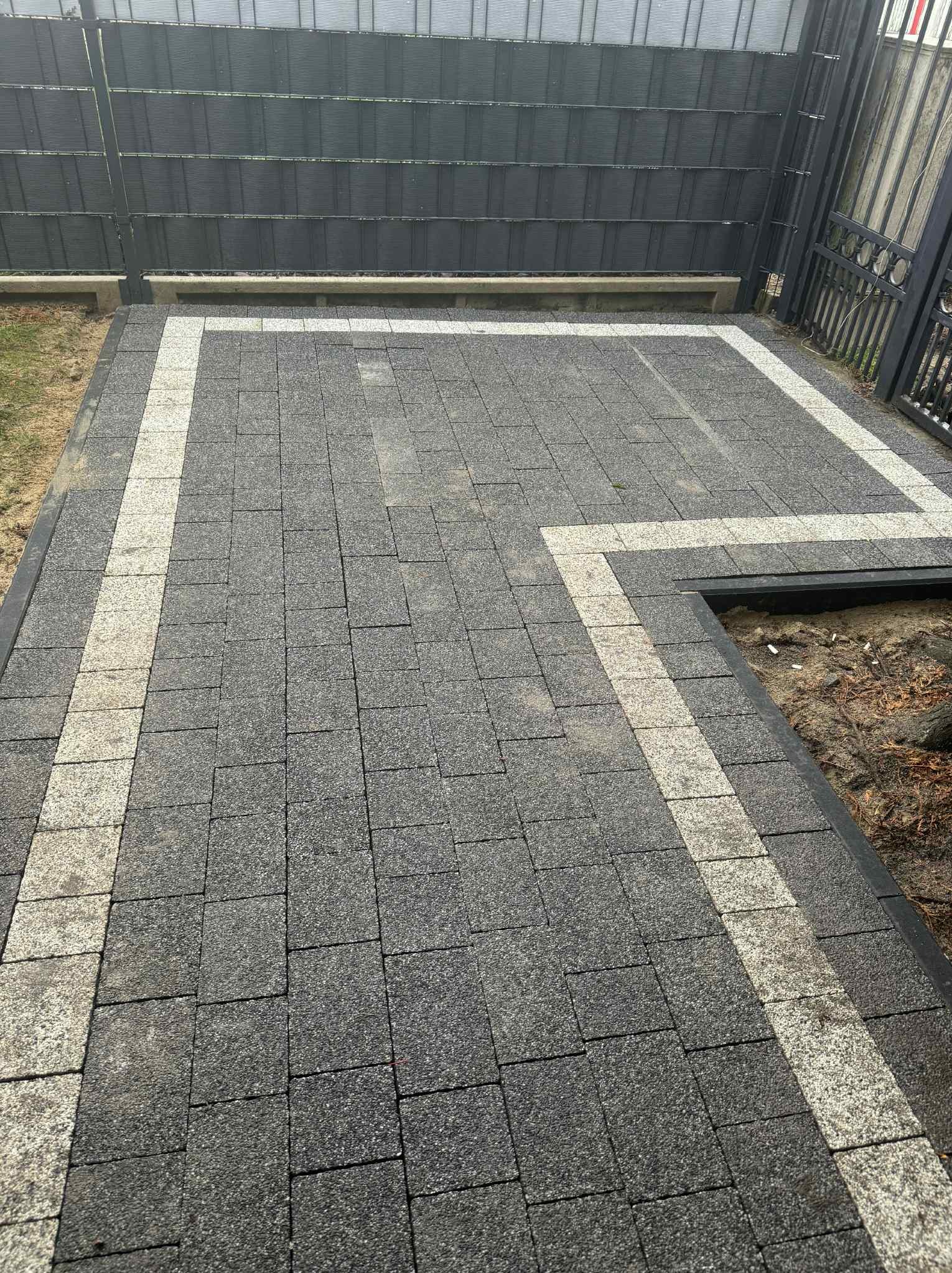 Ułożona szara kostka brukowa z jasną obwódką, widok z góry. Fragment rabaty z obrzeżem. Ciemne ogrodzenie w tle. Regularny wzór kostki.