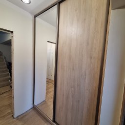 Kamil Meble Na Wymiar - Szafa wnękowa z przesuwnymi drzwiami: lustrzane i drewnopodobne panele. Widoczna podłoga i fragment schodów w tle. Oświetlenie sufitowe.