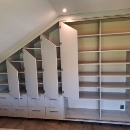 Kamil Meble Na Wymiar - Biała szafa wnękowa z szufladami i półkami, dopasowana do skosu dachu. Część frontów szafek jest otwarta, ukazując puste wnętrze. Widoczne okucia i oświetlenie punktowe.