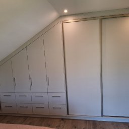 Kamil Meble Na Wymiar - Szafa wnękowa na poddaszu z przesuwnymi drzwiami i szufladami w jasnej kolorystyce, dopasowana do skosu dachu. Widoczne półki po lewej stronie.