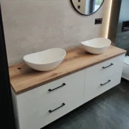 Nowoczesna łazienka z szafką pod umywalki w kolorze białym z drewnianym blatem. Na blacie dwie białe, owalne umywalki nablatowe. W tle szara ściana i okrągłe lustro.