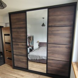 Kamil Meble Na Wymiar - Szafa wnękowa z przesuwnymi drzwiami i lustrem, wykończona w ciemnym drewnie z czarnymi akcentami, odbijająca wnętrze sypialni z łóżkiem i garderobą.