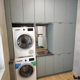 Kamil Meble Na Wymiar - Szara zabudowa meblowa z pralką i suszarką w łazience. Uwagę przykuwa lustro wbudowane w szafkę, odbijające wnętrze łazienki. Drewnopodobna podłoga.