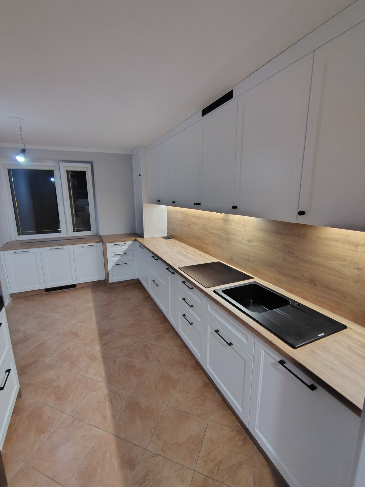 Jasna kuchnia z białymi frontami, drewnianym blatem i czarnymi detalami: uchwyty, zlew i płyta. Podświetlenie blatu dodaje ciepła. Widok na okno.