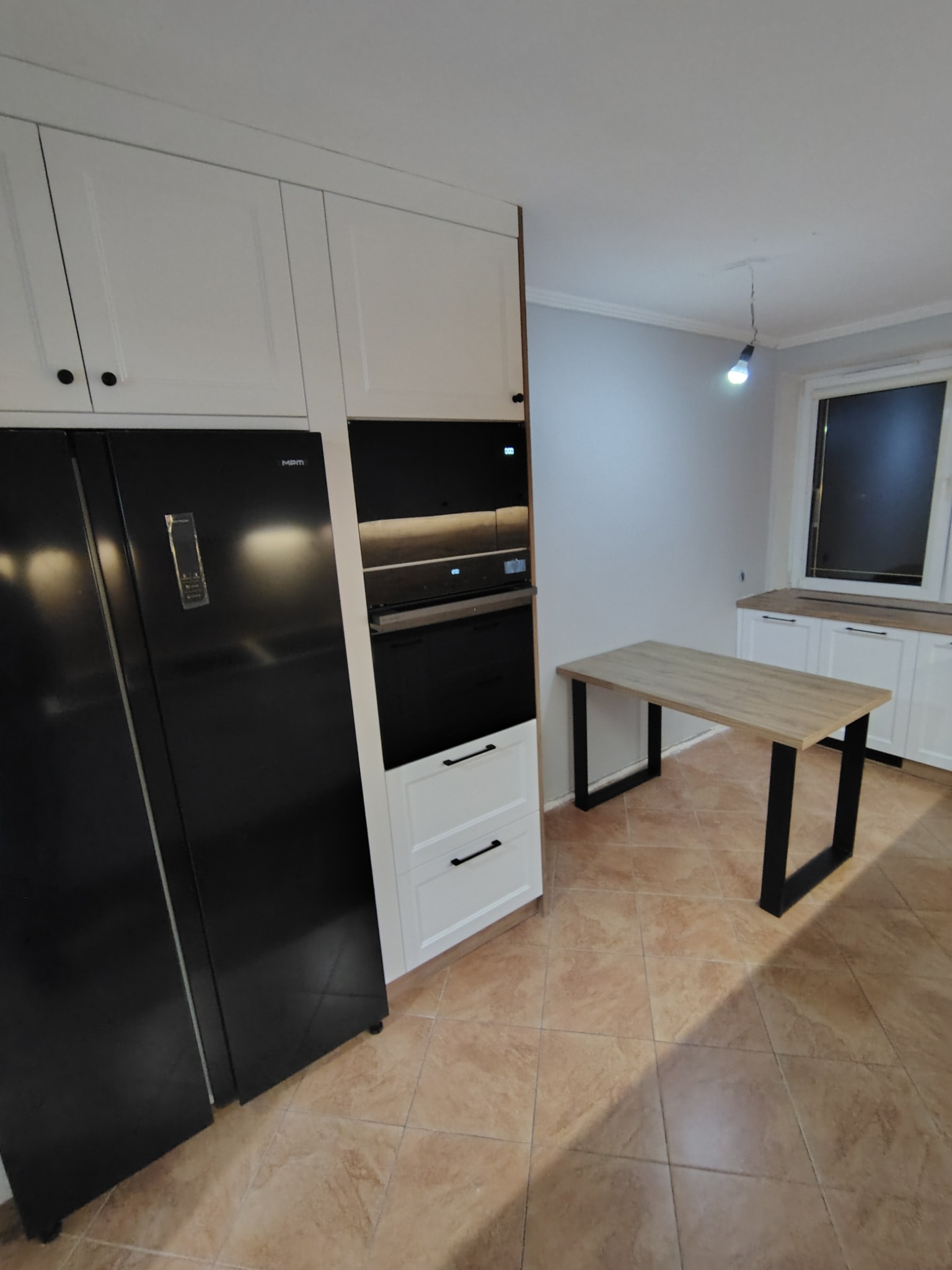 Nowoczesna kuchnia z białymi szafkami, czarną lodówką MPM i stołem z drewnianym blatem na czarnych nogach. Widoczny piekarnik w zabudowie i brązowe płytki podłogowe.