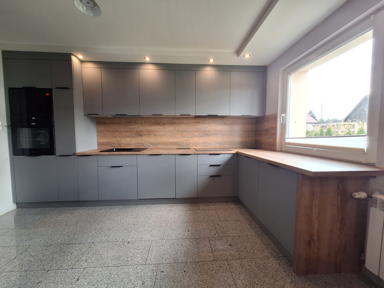 Szara kuchnia z drewnianym blatem i panelem, minimalistyczne uchwyty, wbudowany piekarnik, oświetlenie punktowe, widok na okno i fragment grzejnika.