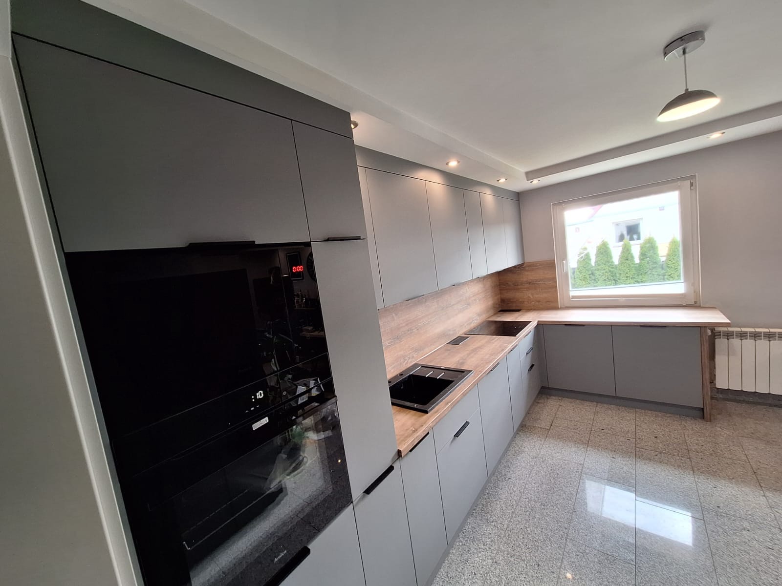 Nowoczesna kuchnia w odcieniach szarości z drewnianym blatem i czarnymi akcentami, piekarnik w zabudowie, widok na okno z zielenią na zewnątrz.