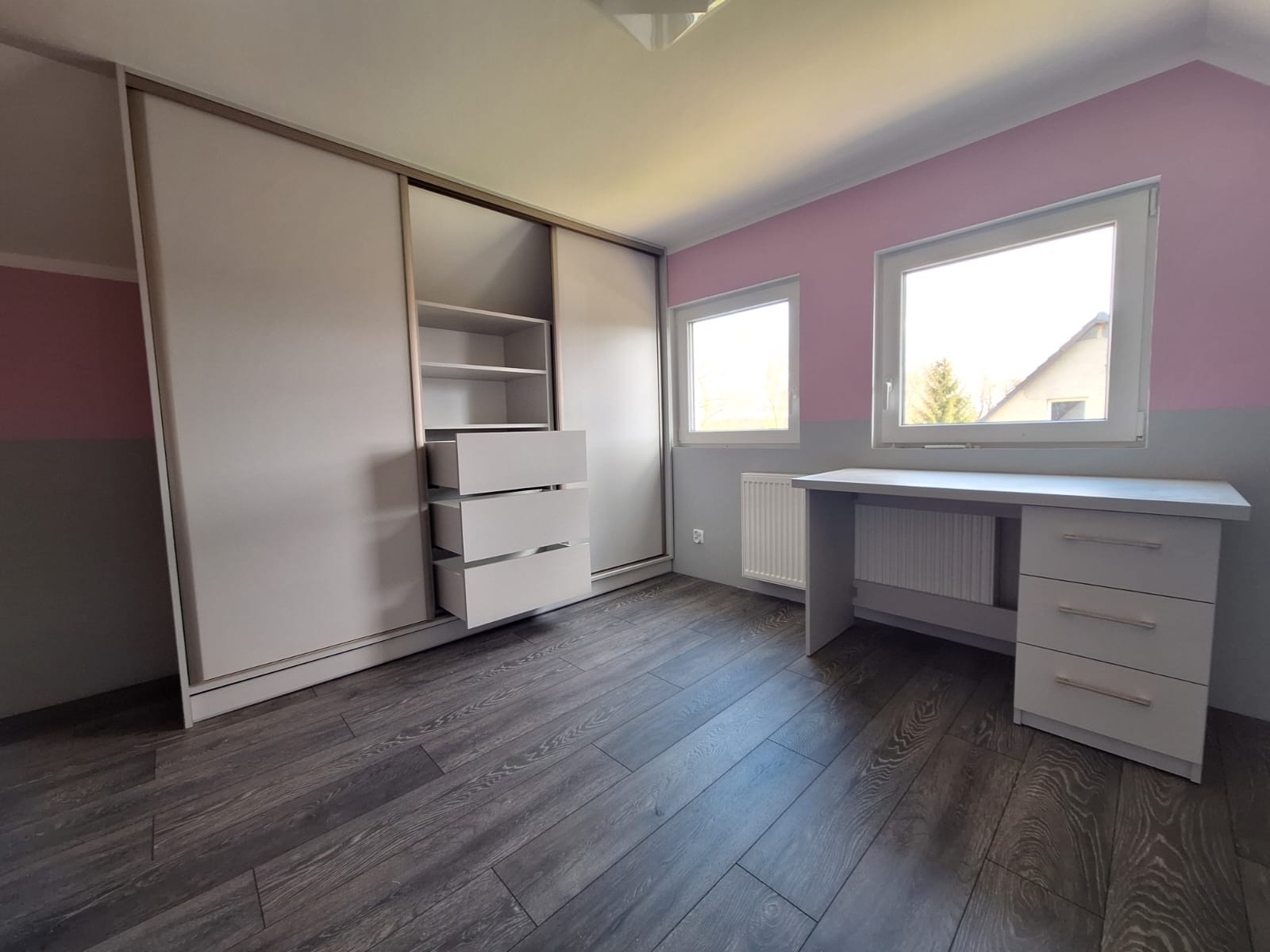 Jasna szafa przesuwna z otwartymi szufladami i biurko w pokoju z panelami podłogowymi i ścianami w kolorze różowo-szarym, oświetlonym naturalnym światłem z okien.