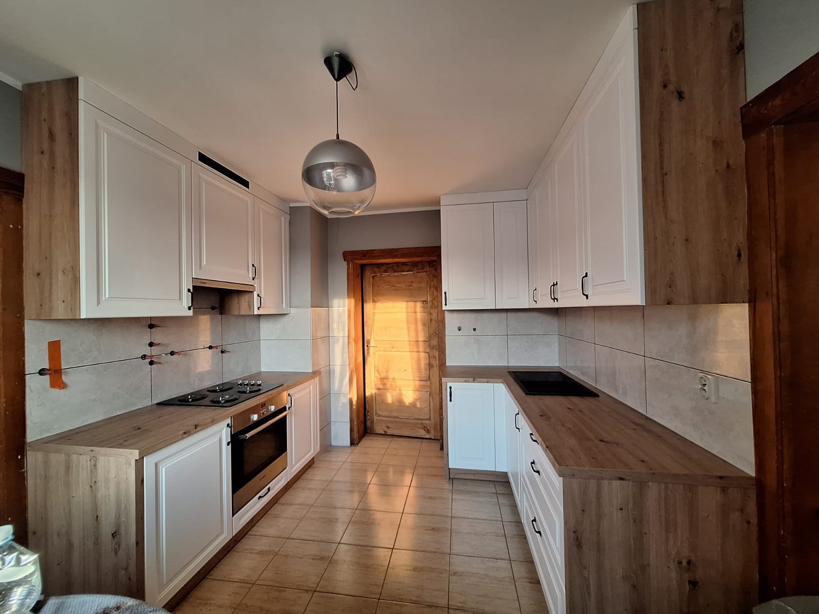 Jasna kuchnia z białymi szafkami i drewnianymi blatami, oświetlona kulistą lampą. Na ścianie widoczne krzyżyki dystansowe do płytek.