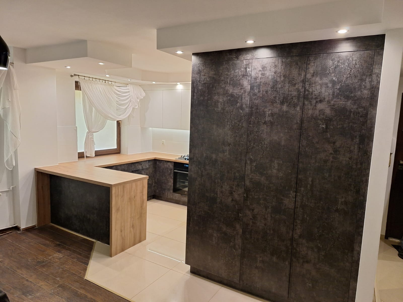 Nowoczesna kuchnia z wyspą i zabudową szaf w kolorze grafitowym, jasne blaty, białe szafki wiszące, okno z firaną. Ukończona realizacja.