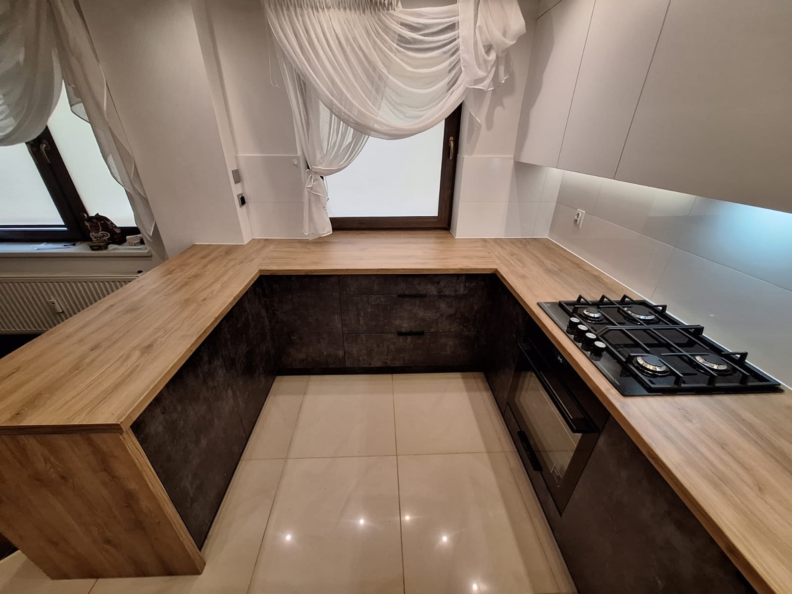 Nowoczesna kuchnia w kształcie litery U: drewniany blat, szare szafki z ciemnym wzorem, okno z firanką, płyta gazowa. Ujęcie z perspektywy podłogi.