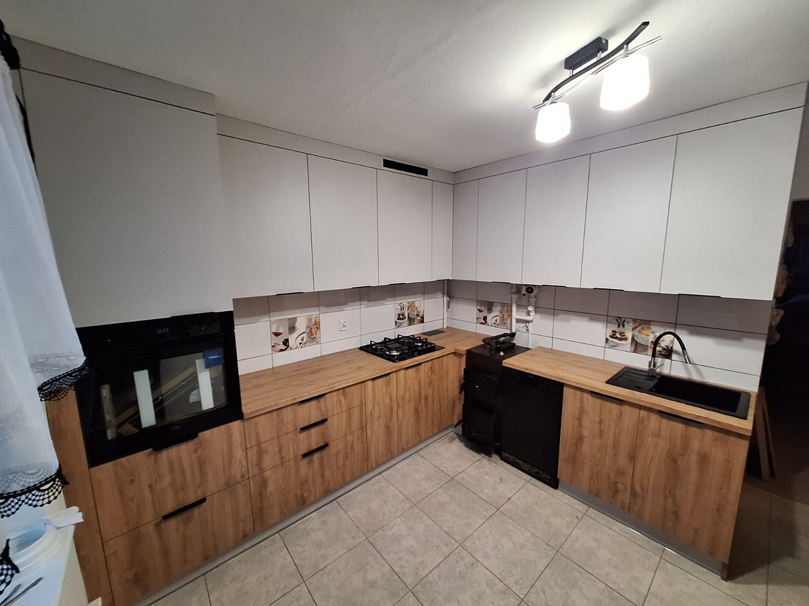 Nowoczesna kuchnia z białymi górnymi szafkami i drewnopodobnymi dolnymi, czarny piekarnik i płyta, płytki z motywem kawy, oświetlenie sufitowe.