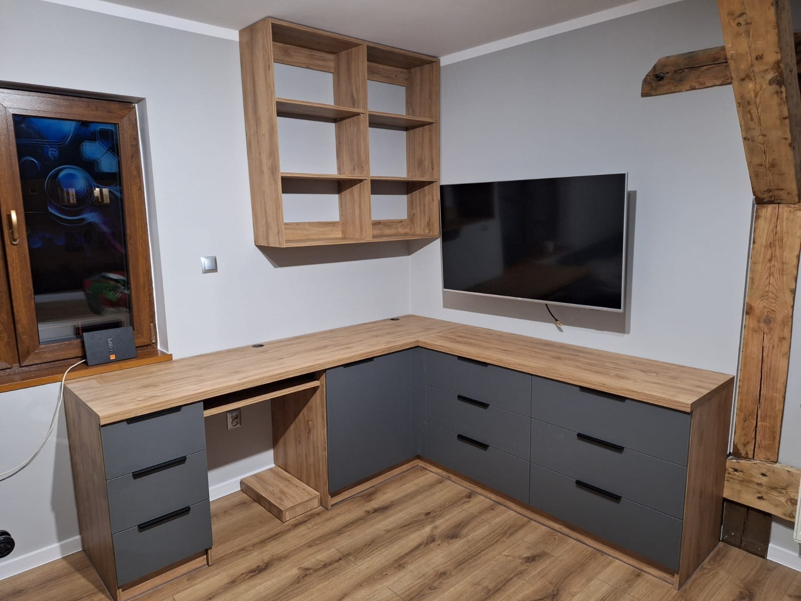 Drewniane biurko na wymiar z szufladami w szarym kolorze, nad nim półki i telewizor. Widoczny fragment drewnianej belki. Aranżacja w szarościach i drewnie.