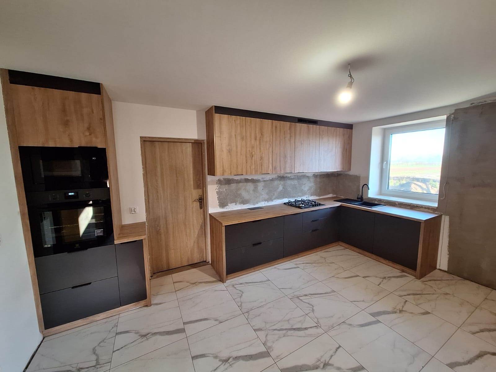 Nowoczesna kuchnia z szarymi szafkami dolnymi i drewnopodobnymi górnymi, czarnym sprzętem AGD i marmurową podłogą w trakcie wykańczania.