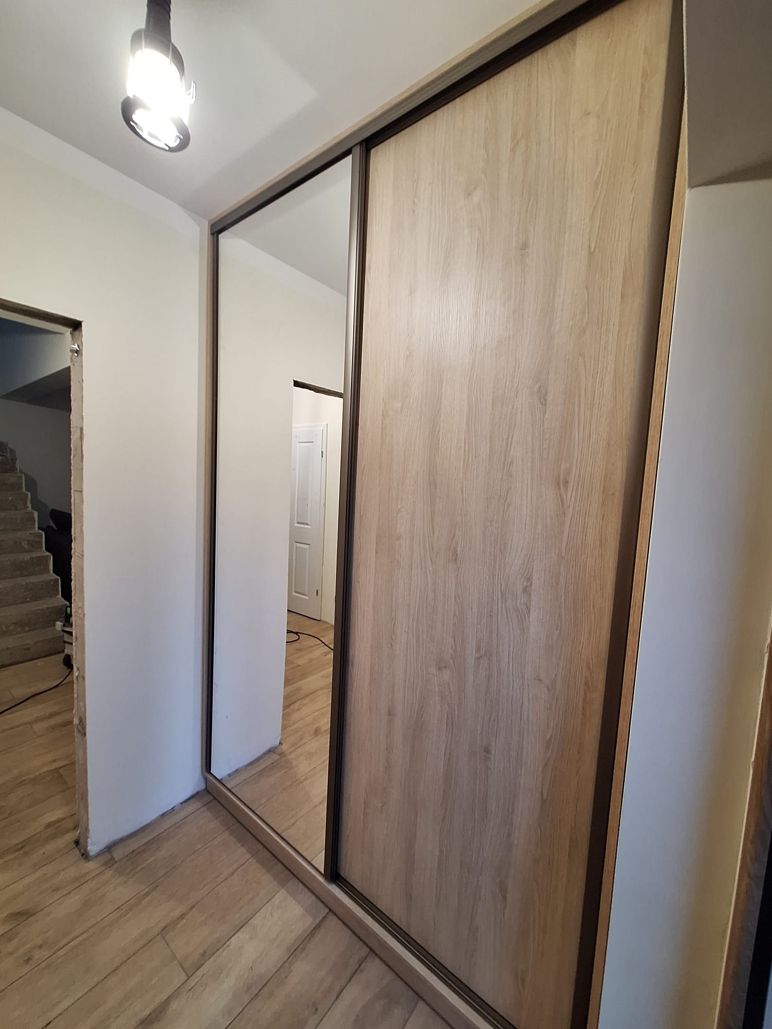 Szafa wnękowa z przesuwnymi drzwiami: lustrzane i drewnopodobne panele. Widoczna podłoga i fragment schodów w tle. Oświetlenie sufitowe.