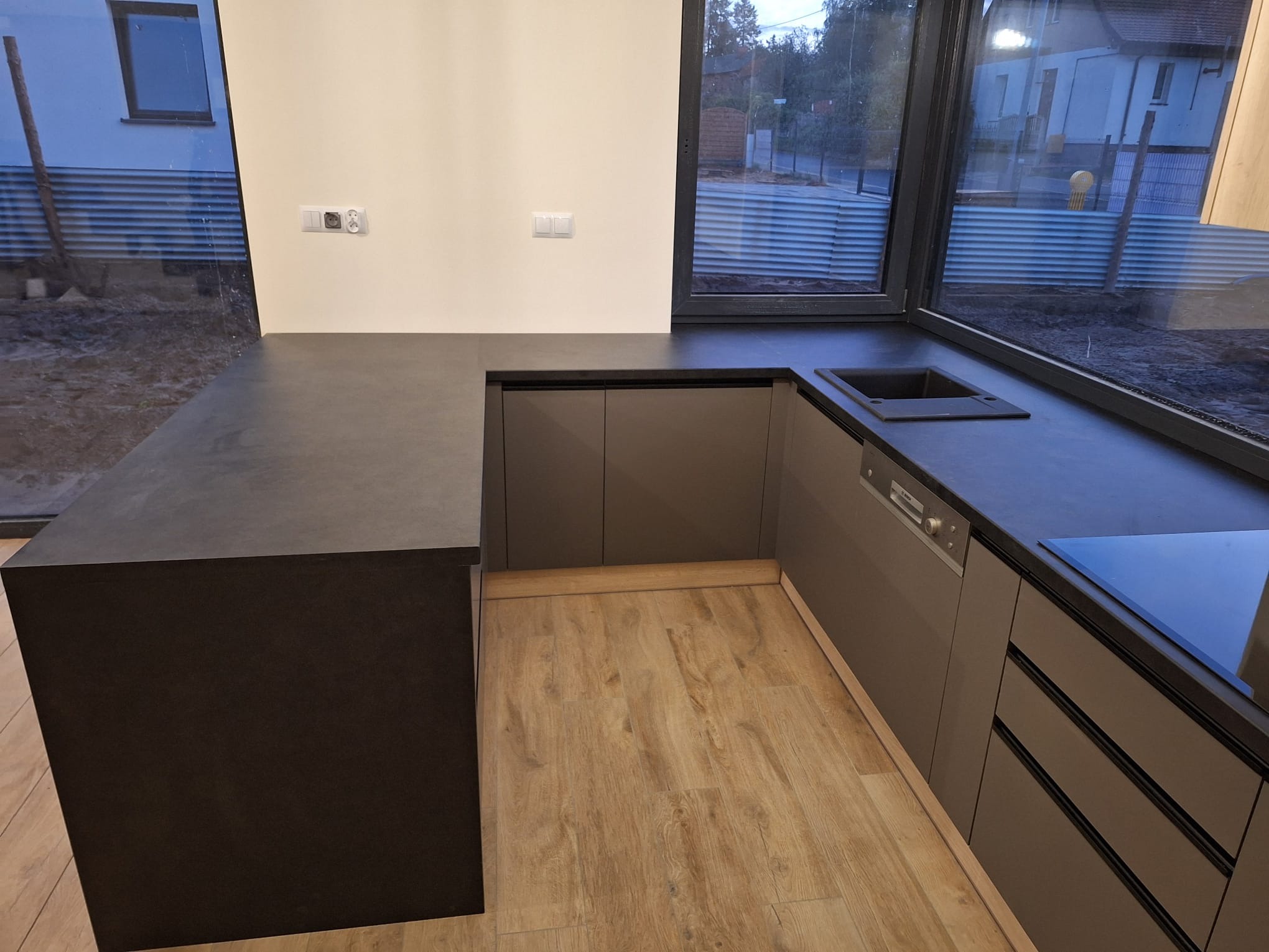 Nowoczesna kuchnia z ciemnoszarym blatem i frontami, drewnopodobna podłoga, okno z widokiem na zewnątrz. Minimalistyczny design z elementami zabudowy.