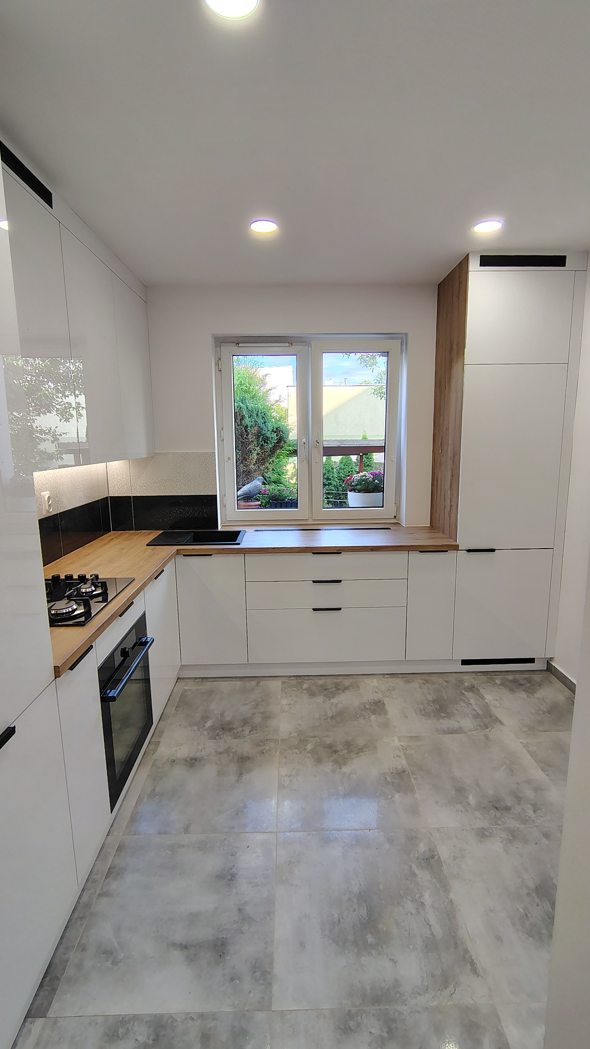 Nowoczesna, biała kuchnia na wymiar z drewnianym blatem w Starym Gronowie. Widok na ogród przez okno, minimalistyczne uchwyty.