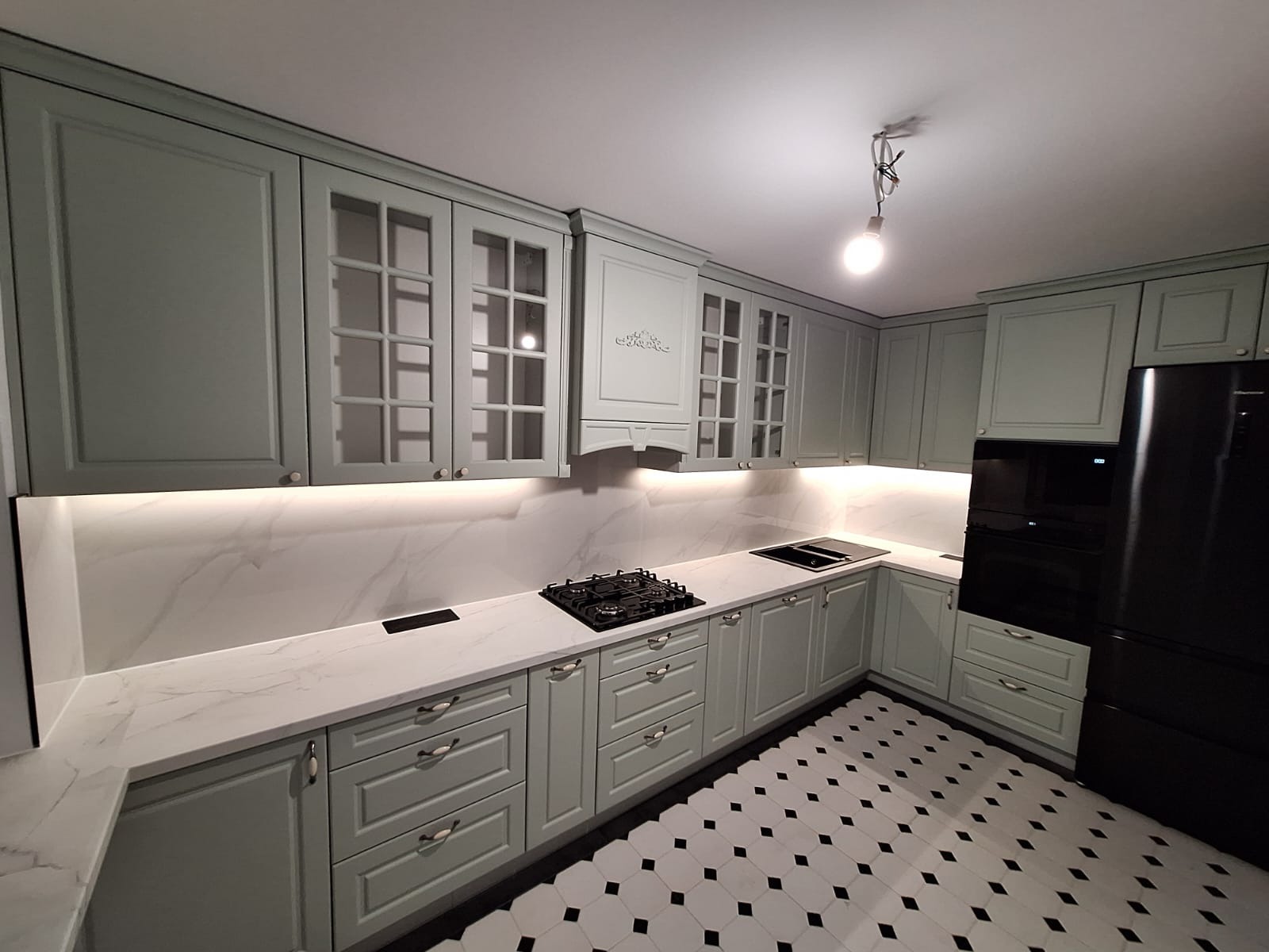 Jasnozielona kuchnia z frezowanymi frontami, szklanymi witrynami i marmurowym blatem. Czarno-biała podłoga w romby i podświetlenie LED pod szafkami.