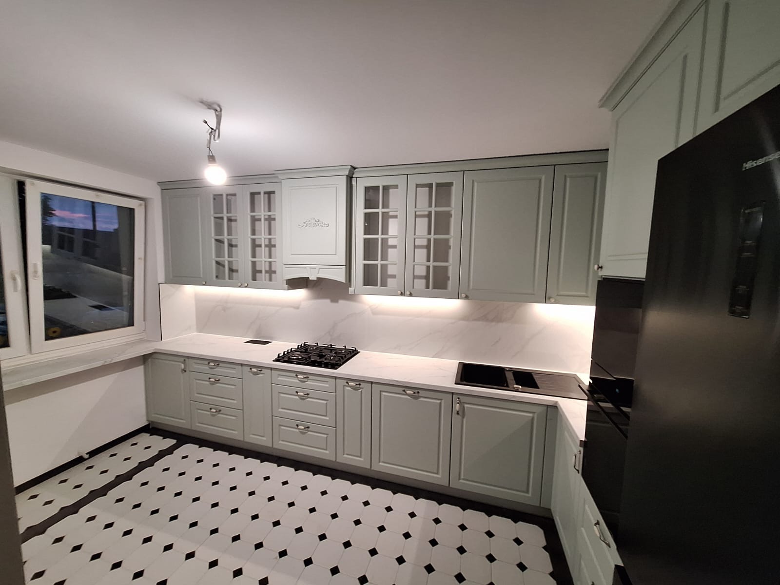 Jasna kuchnia z frezowanymi frontami, marmurowym blatem i czarno-białą podłogą w romby. Uwagę zwraca dekoracyjny okap i przeszklone szafki wiszące.