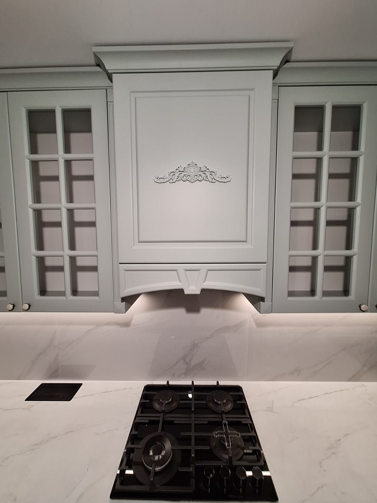 Elegancka zabudowa kuchenna w odcieniach szarości z dekoracyjnym ornamentem nad płytą gazową. Szafki z przeszklonymi frontami i subtelnym oświetleniem blatu.