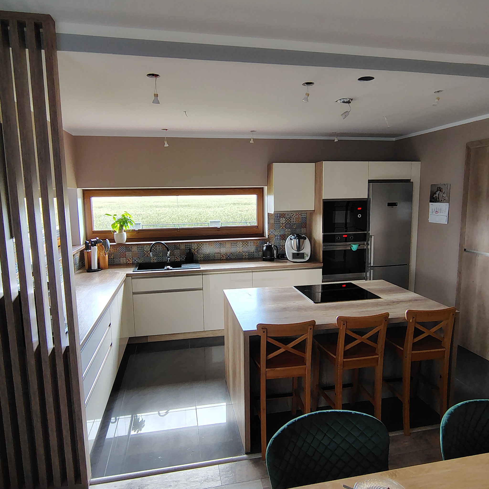 Jasna kuchnia z wyspą i drewnianymi hokerami. Nad blatem okno z widokiem na zieleń. Kolorowe płytki nad blatem. Widoczne niedokończone oświetlenie sufitowe.