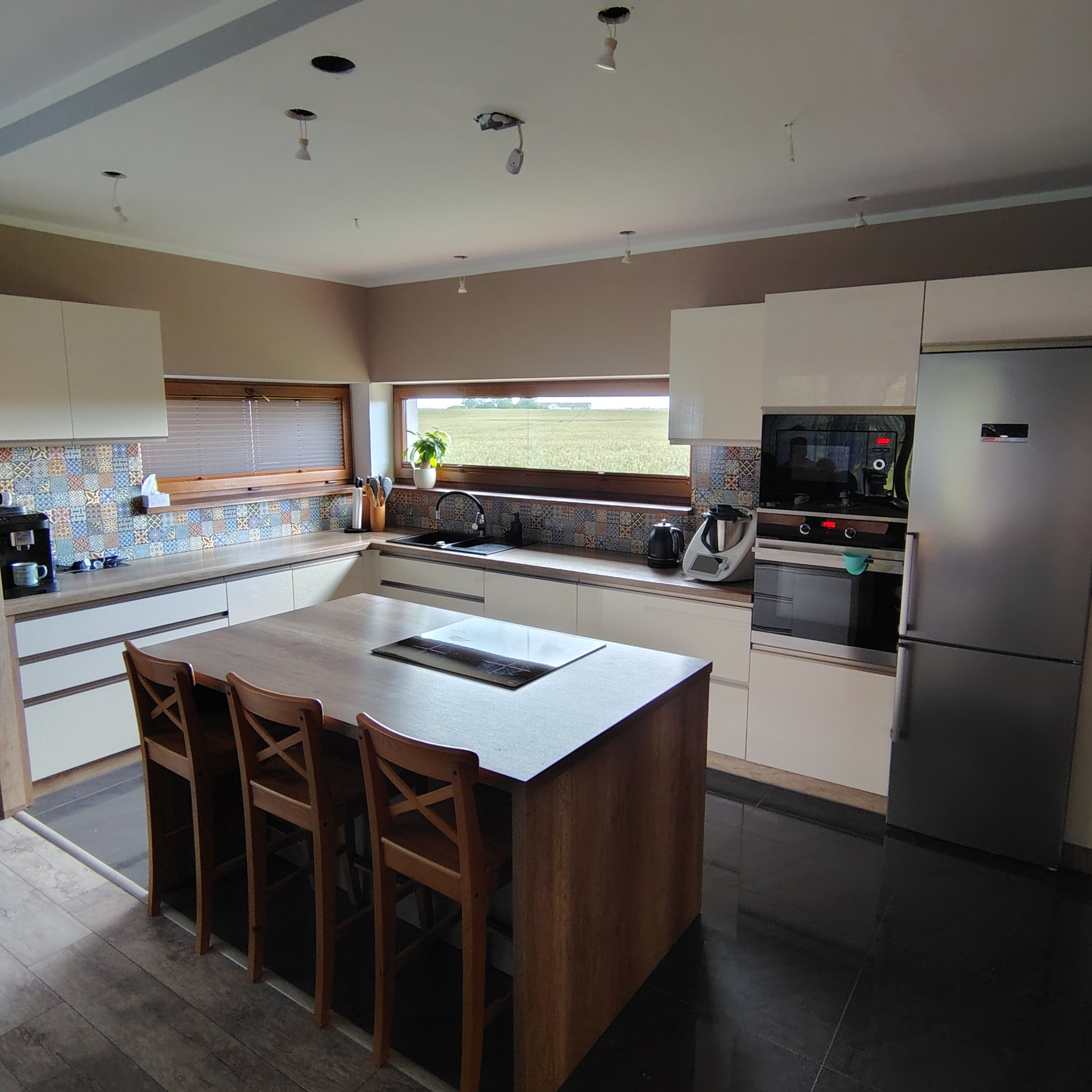 Jasna kuchnia z wyspą i drewnianymi hokerami. Nad blatem okno z widokiem na pole. Kolorowe płytki nad blatem. Częściowo widoczne niedokończone oświetlenie.