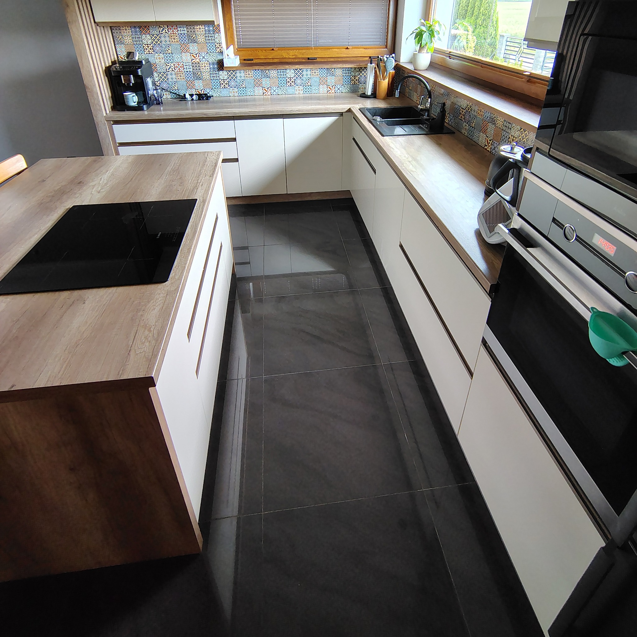 Nowoczesna kuchnia z białymi szafkami, drewnianym blatem i wyspą z płytą indukcyjną. Czarne płytki podłogowe. Widok na okno i akcesoria kuchenne.