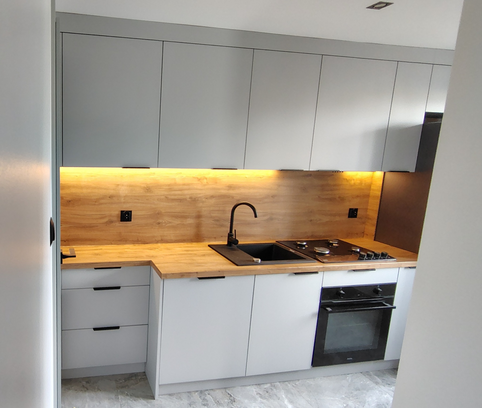 Nowoczesna kuchnia z szarymi szafkami górnymi i białymi dolnymi, drewnianym blatem i podświetleniem LED. Ujęcie z perspektywy wejścia do pomieszczenia.