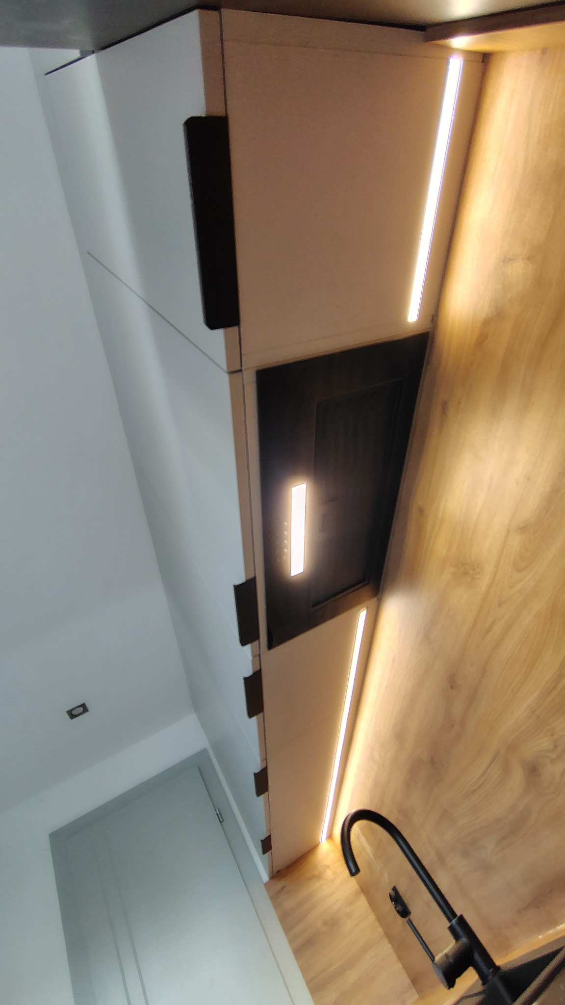 Nowoczesna kuchnia z białymi szafkami, czarnymi uchwytami i drewnianym blatem. Podświetlenie LED podkreśla minimalistyczny design i dodaje elegancji.