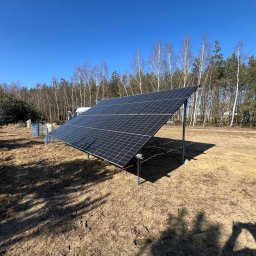 TERMOECO SPÓŁKA Z OGRANICZONĄ ODPOWIEDZIALNOŚCIĄ - Nowoczesna instalacja fotowoltaiczna na gruncie, panele ustawione pod kątem, widoczne mocowania, w tle drzewa i błękitne niebo.