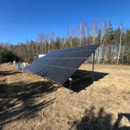 Nowoczesna instalacja fotowoltaiczna na gruncie, panele ustawione pod kątem, widoczne mocowania, w tle drzewa i błękitne niebo.