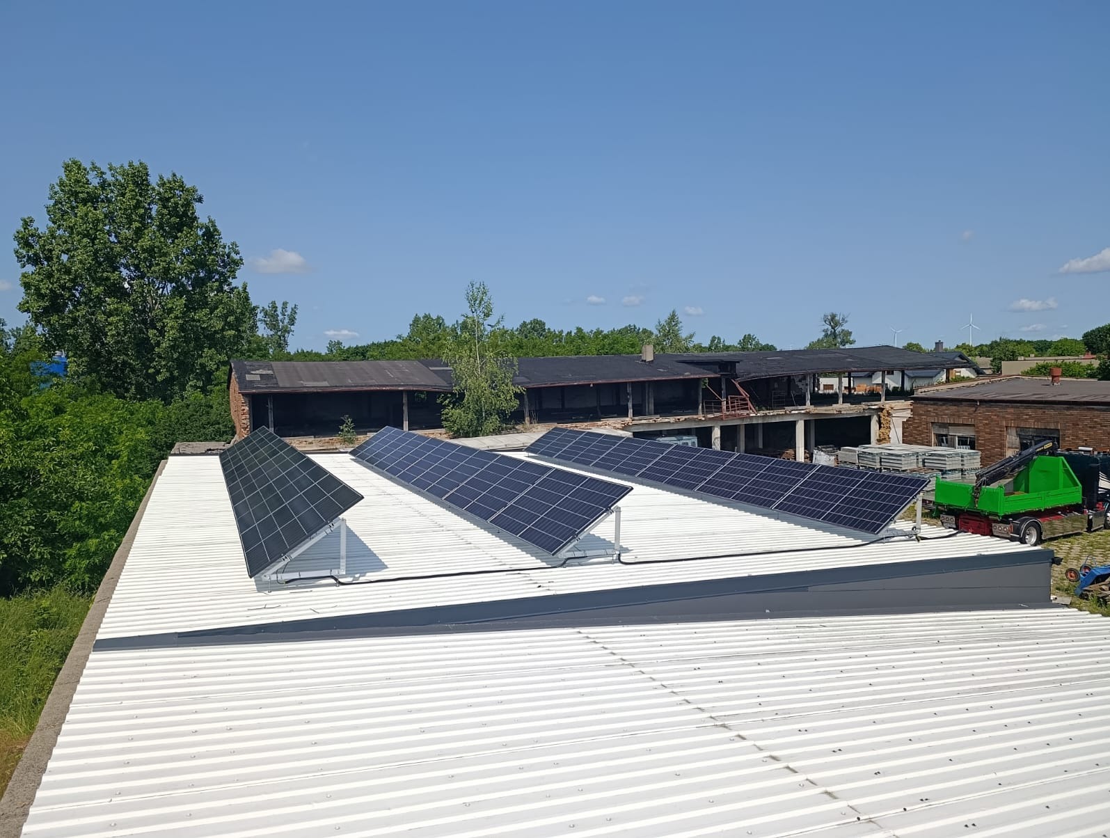 Rząd paneli fotowoltaicznych na białym, trapezowym dachu budynku przemysłowego, w tle zieleń i częściowo zniszczony budynek. Widok z góry, słoneczny dzień.
