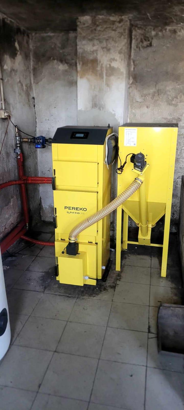 Żółty piec na paliwo stałe Pereko Q-Pell EVO z podajnikiem w kotłowni o surowych ścianach. Widoczne rury instalacji CO.
