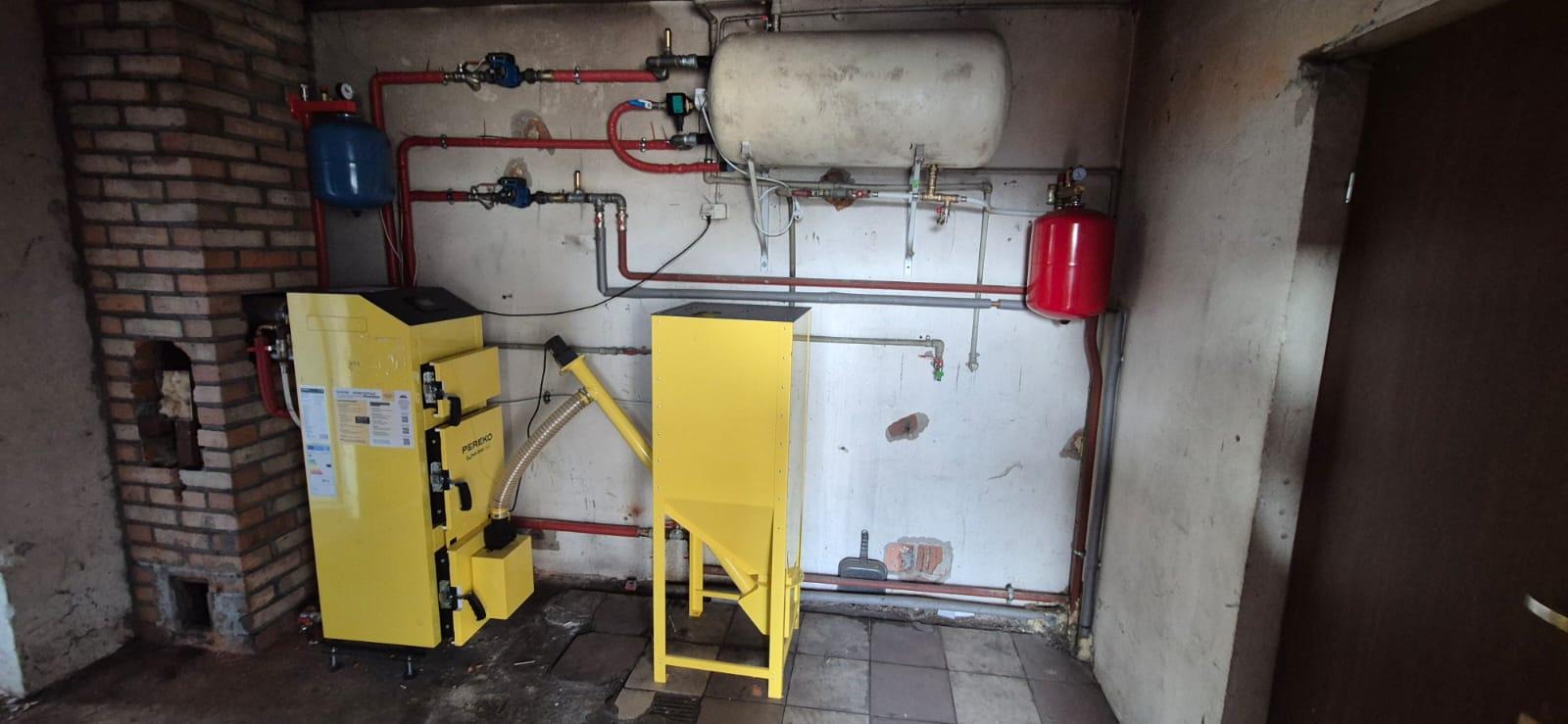 Żółty piec na paliwo stałe Pereko z podajnikiem w kotłowni z instalacją grzewczą i zbiornikami, widok z przodu.