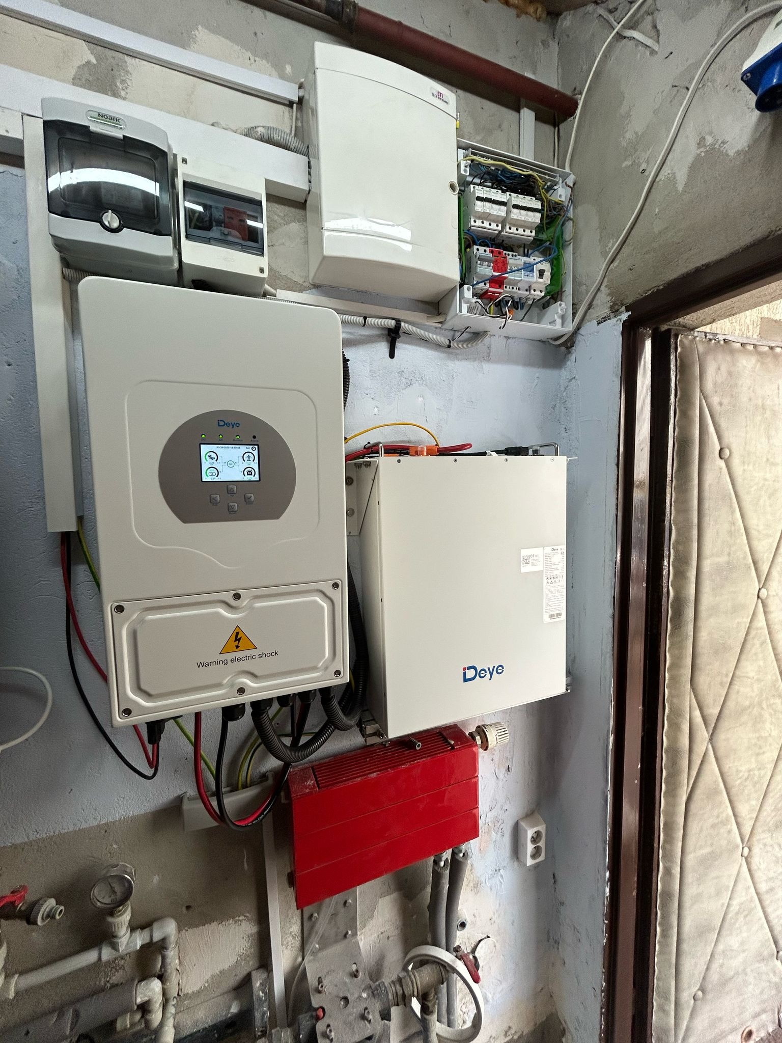 Instalacja fotowoltaiczna: falownik Deye z wyświetlaczem, skrzynka z bezpiecznikami i akumulator na szarej ścianie w pomieszczeniu gospodarczym.