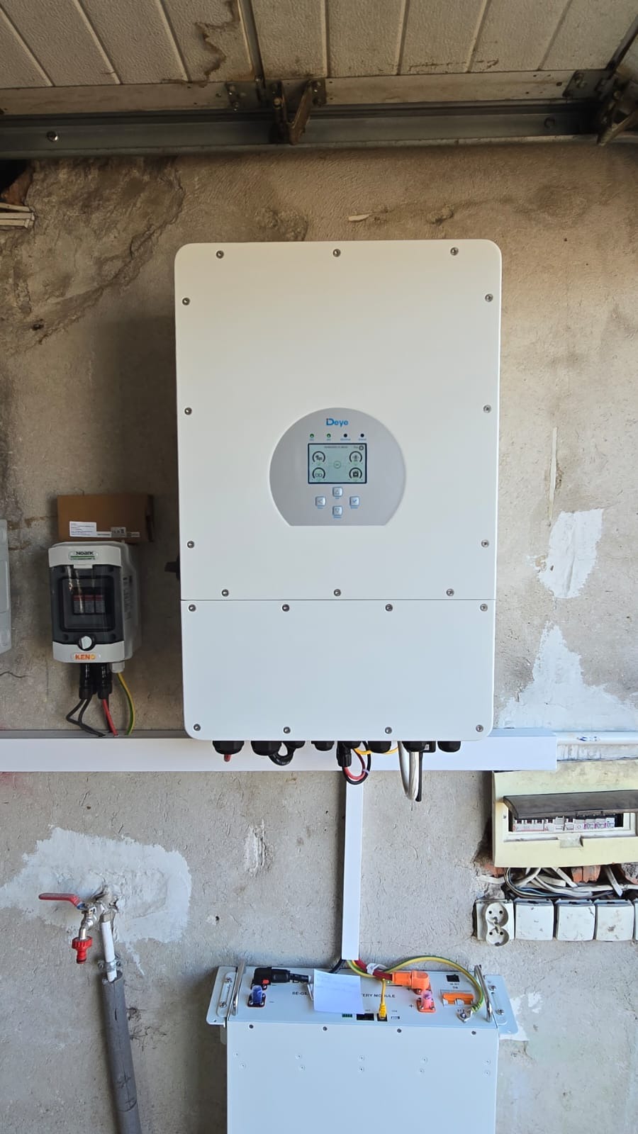 Biały inwerter fotowoltaiczny Deye zamontowany na szarej, surowej ścianie. Widoczne okablowanie, skrzynka z bezpiecznikami i bateria. Surowy beton w tle.