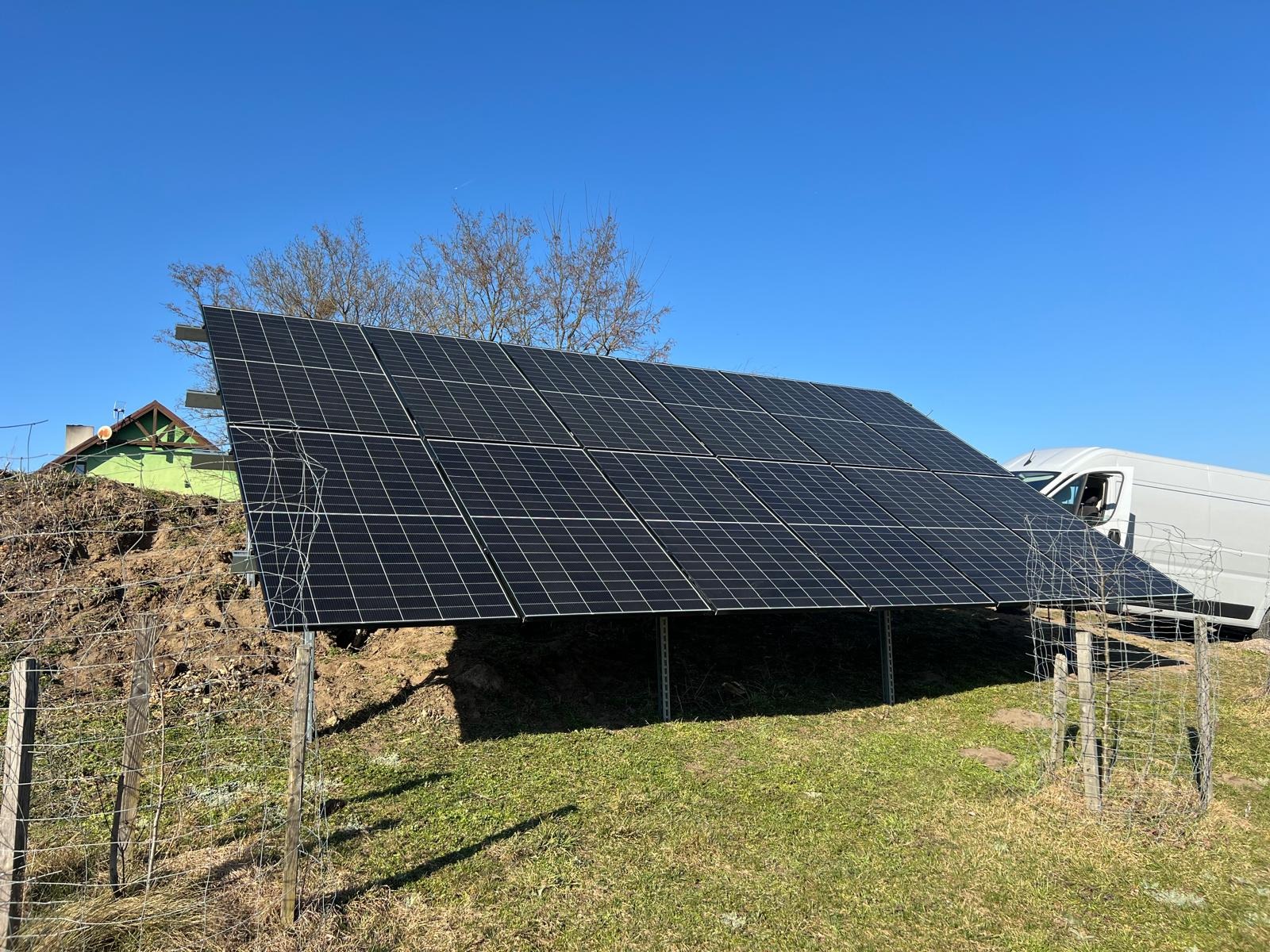 Instalacja fotowoltaiczna na trawiastym terenie w Poznaniu, widoczny biały bus w tle. Panele ustawione pod kątem, błękitne niebo.