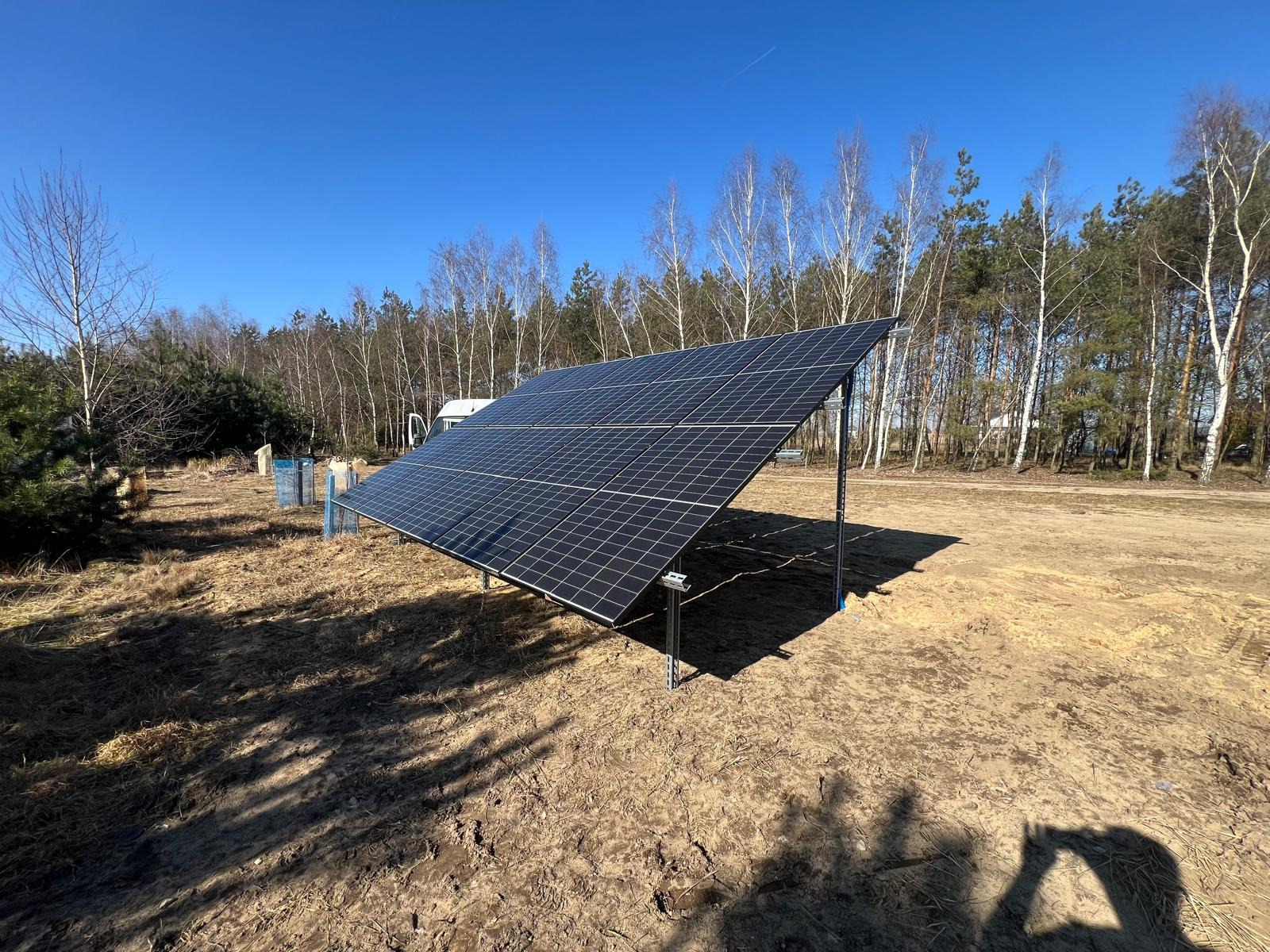 Nowoczesna instalacja fotowoltaiczna na gruncie, panele ustawione pod kątem, widoczne mocowania, w tle drzewa i błękitne niebo.
