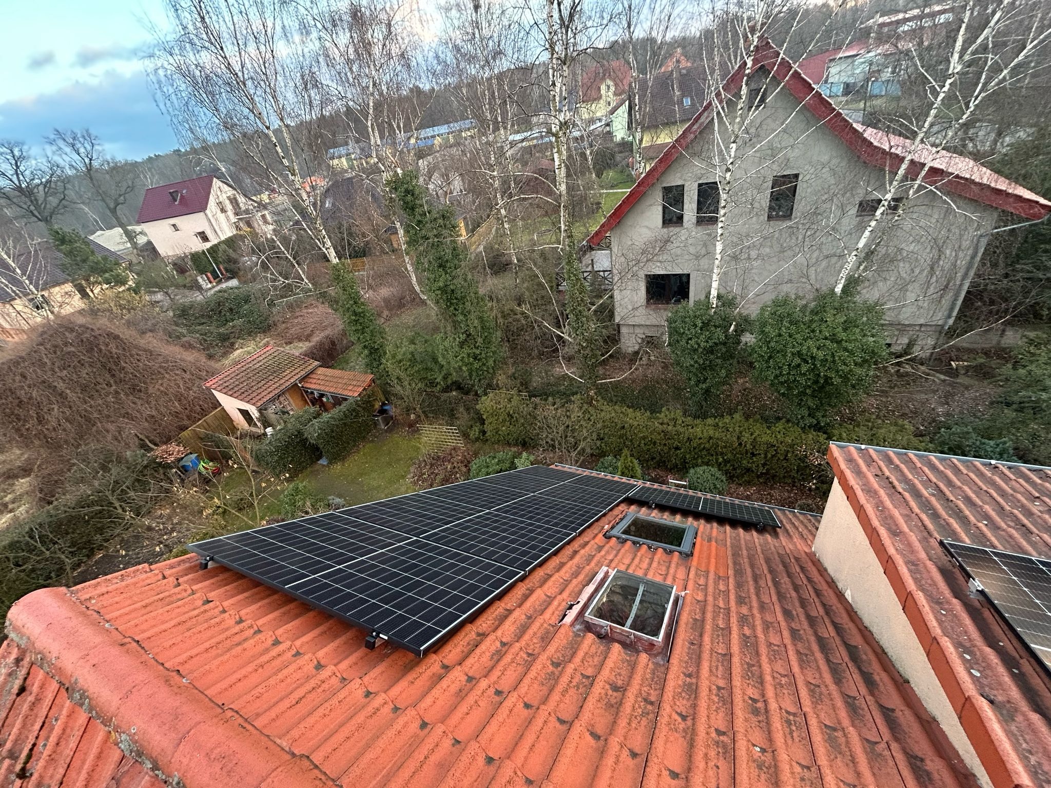 Panele słoneczne zamontowane na dachu z czerwonej dachówki, widok z góry na tle drzew i zabudowań. Widoczne okna dachowe.