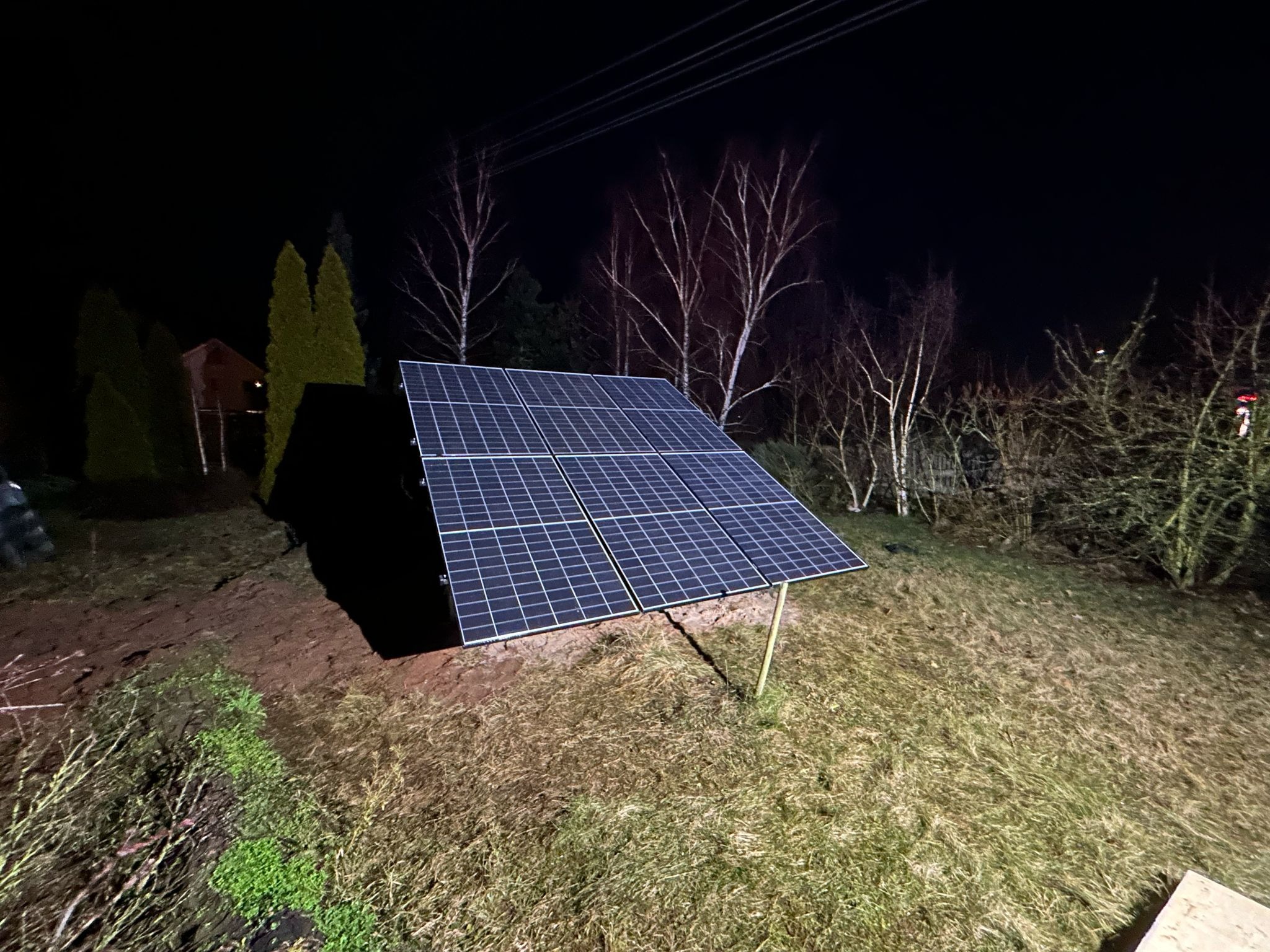 Nocny widok na panele fotowoltaiczne ustawione na trawiastym wzgórzu, z drzewami w tle i ciemnym niebem. Widoczne kable elektryczne.