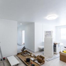 Marcin Czapelka - Jasne wnętrze w trakcie remontu: drabina, narzędzia, materiały budowlane, okno z widokiem na zewnątrz. Widoczne ślady świeżego malowania ścian.