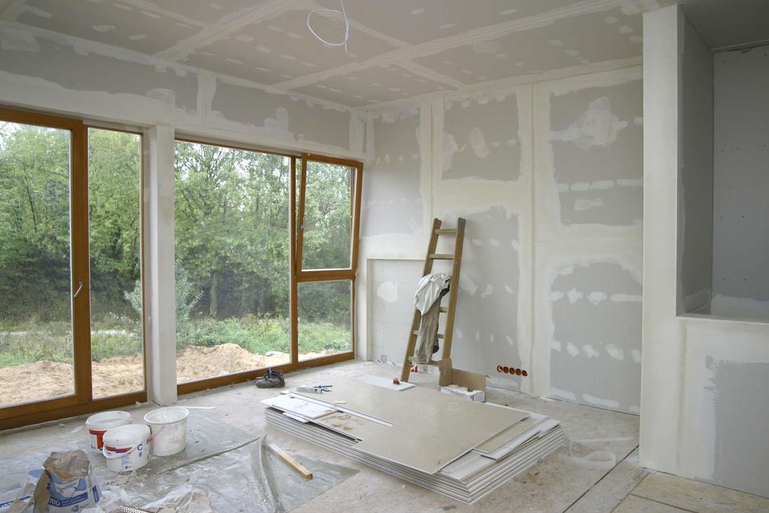 Wnętrze w trakcie remontu: ściany z płyt gipsowo-kartonowych, szpachlowane łączenia, okno z widokiem na zieleń, drabina oparta o ścianę, płyty na podłodze.